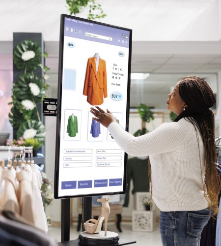 Woman buyer selecting clothes on board, using interactive touch screen monitor in clothing store. Young boutique customer buying fashion items from self ordering kiosk service, retail shop.